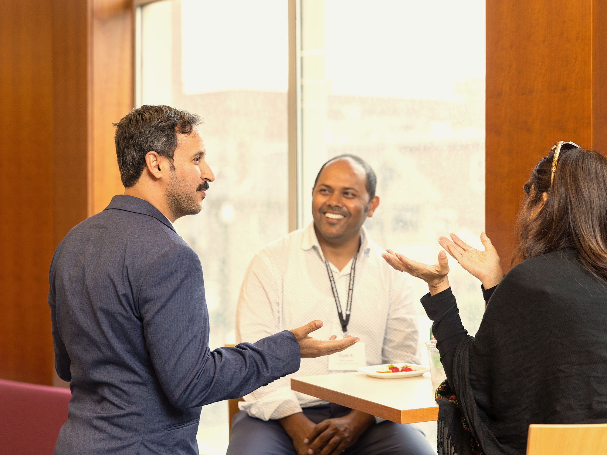Three colleagues engaged in a friendly discussion at a high-top table near a window.