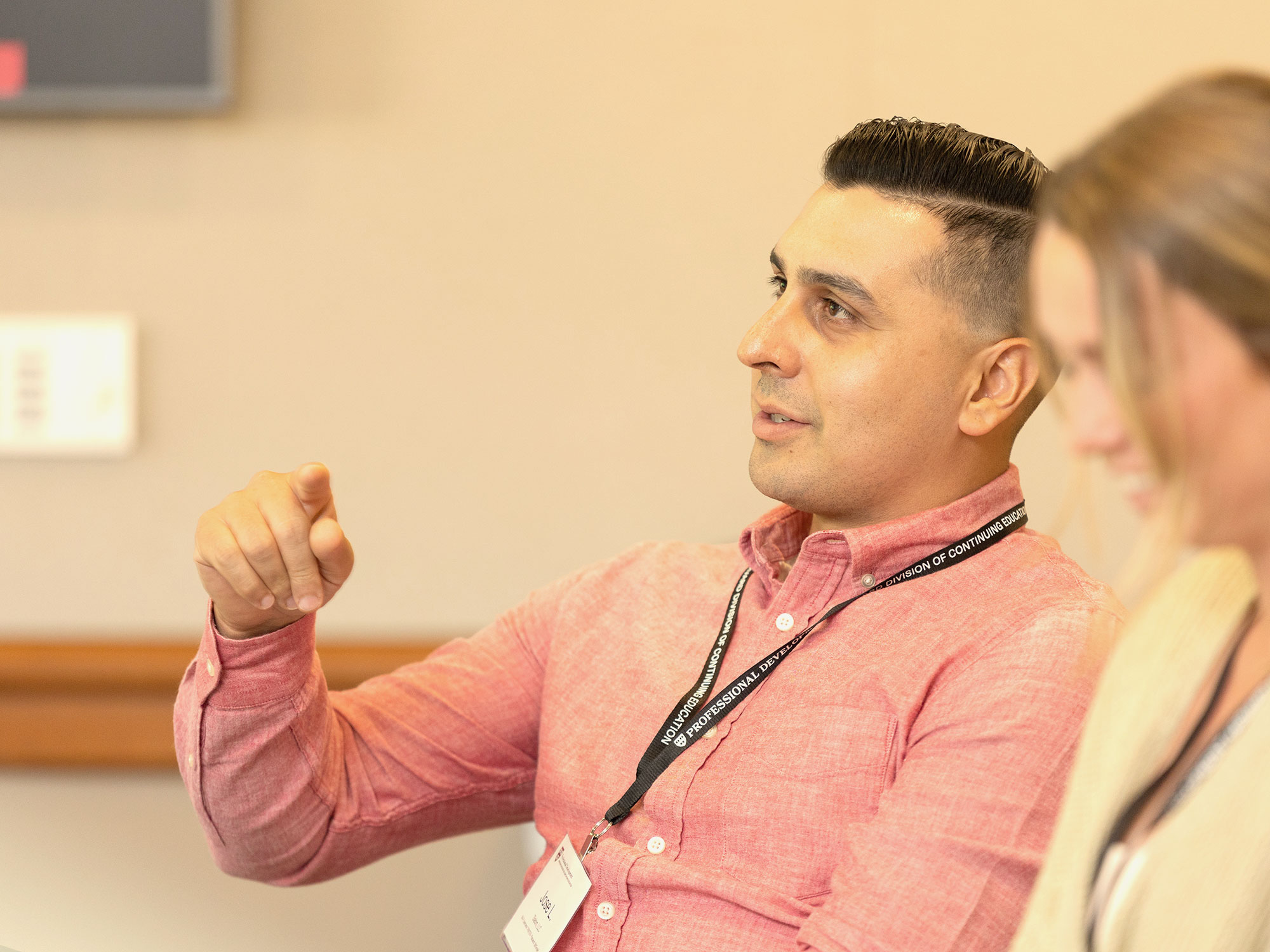 A man in a pink dress shirt speaking while gesturing with his hands. An out of focus woman smiles besides him.
