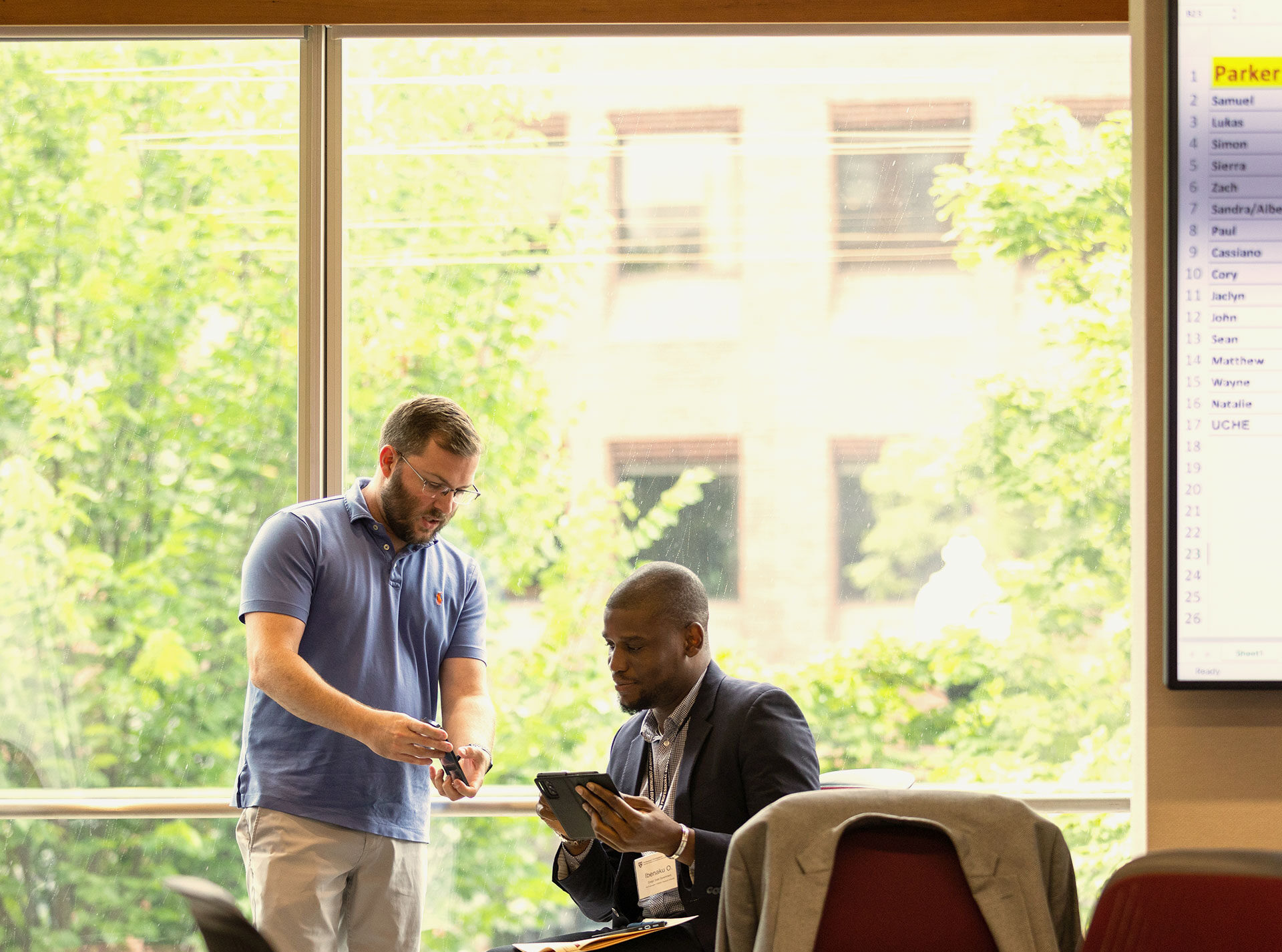 Two men discussing something on a tablet near a large window with greenery outside.