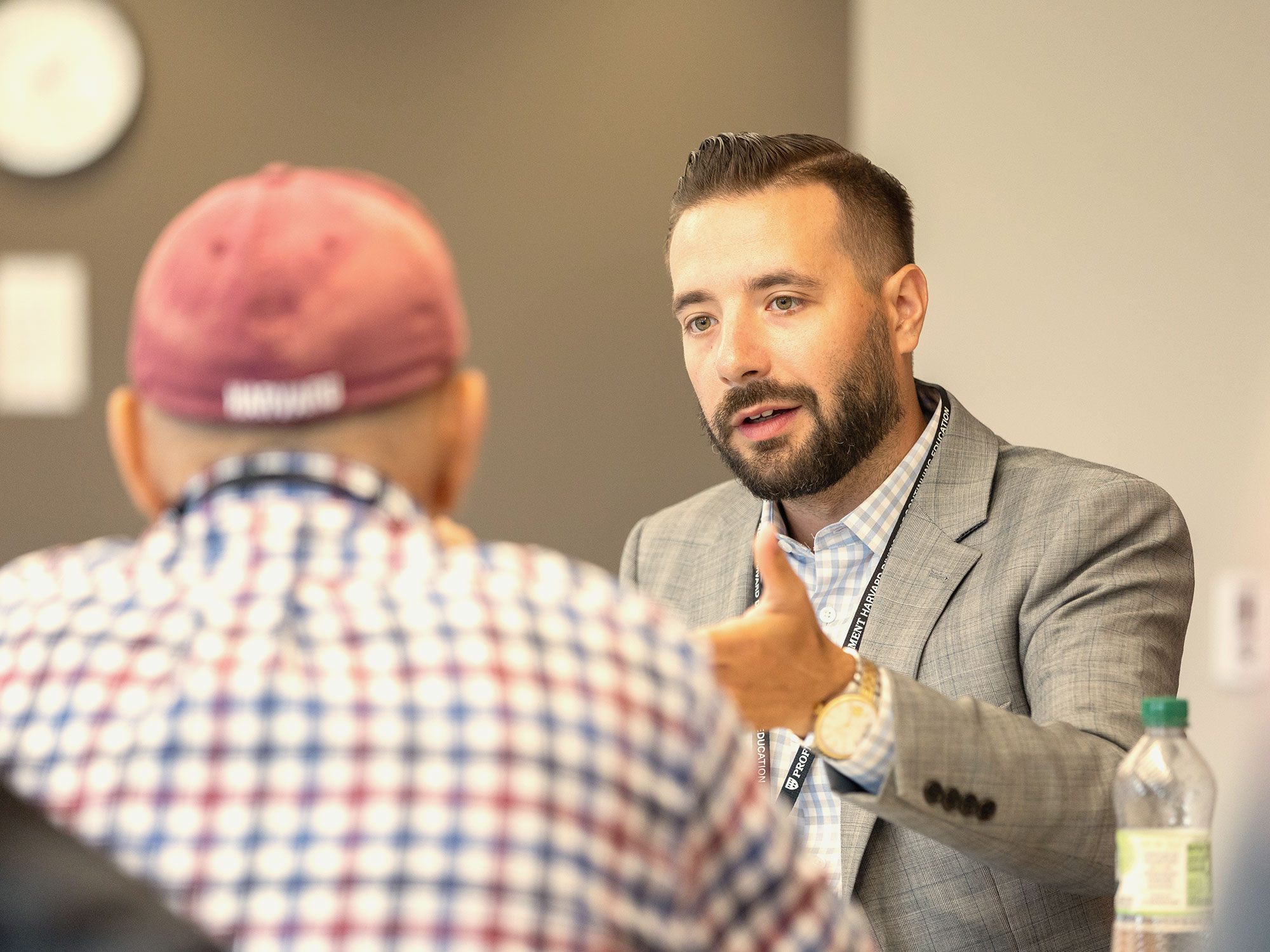 A younger man with a beard in a grey suit speakers to an older man with a plaid shirt and a baseball cap.